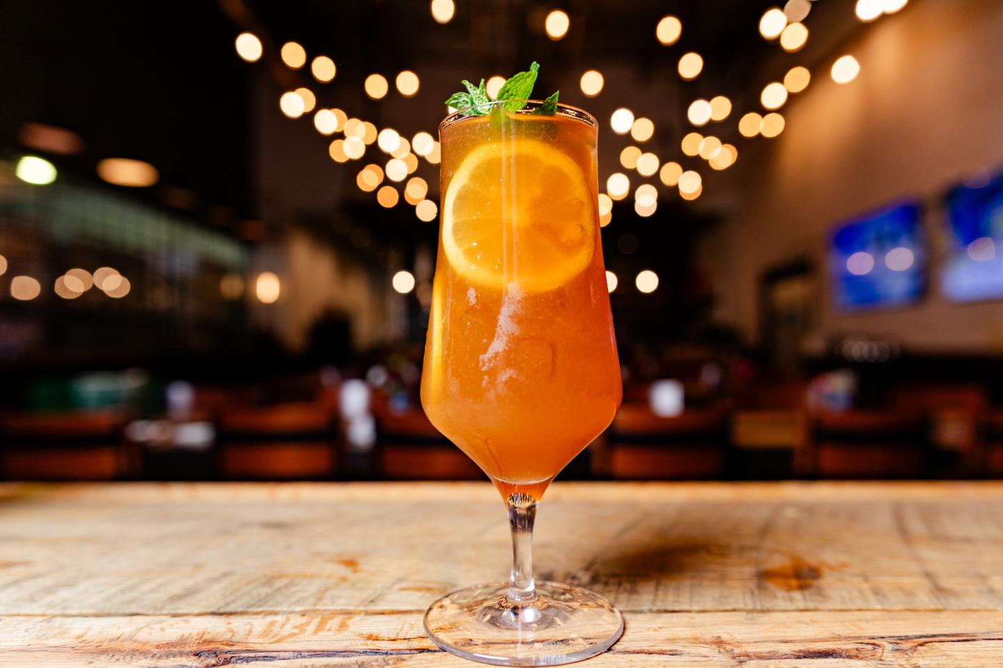 Orange cocktail with mint and orange slice garnish on a wooden table, blurred lights in background.