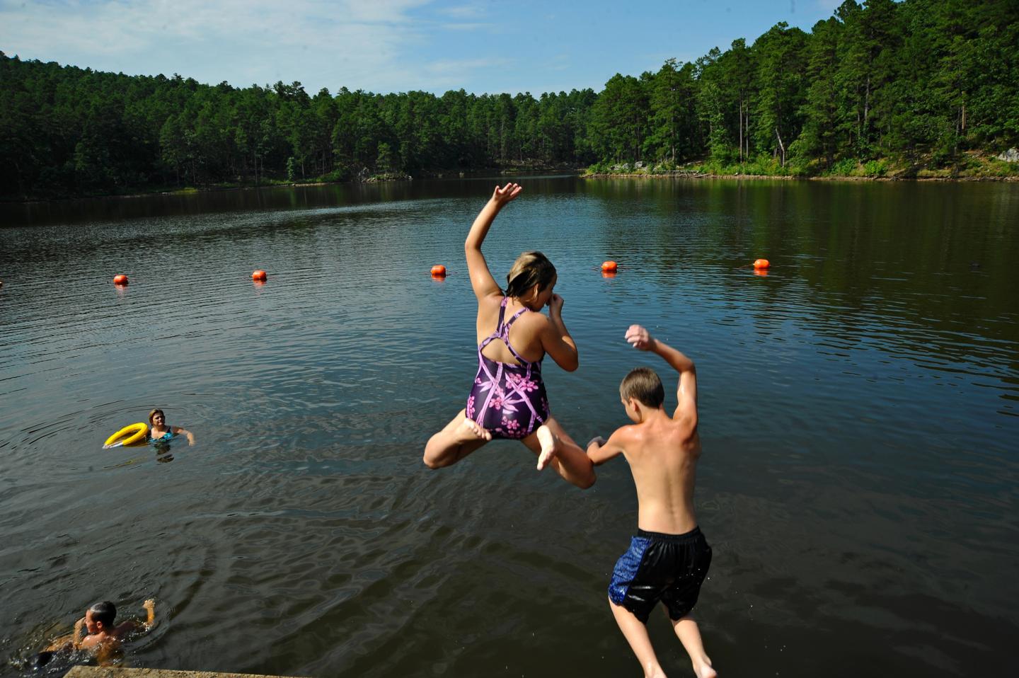 Two people jump into a lake on a sunny day, surrounded by trees and a swimmer.