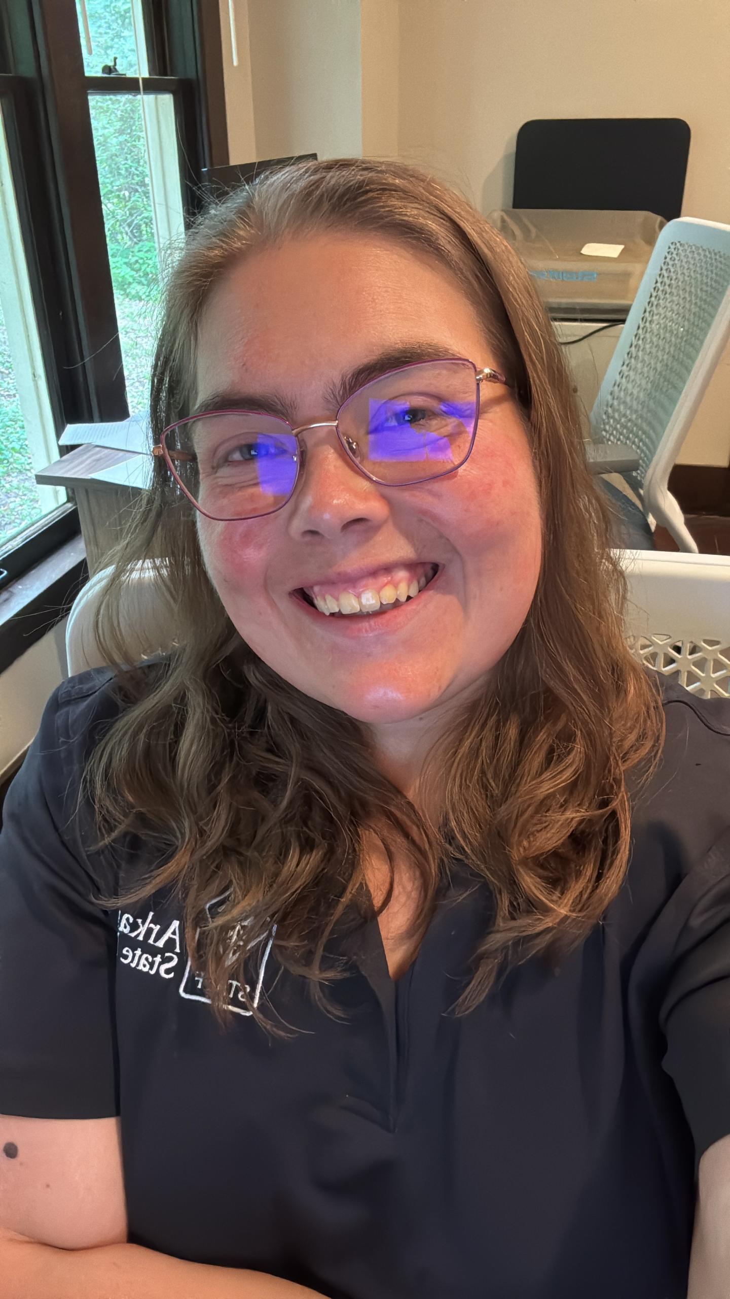 Smiling person with glasses sitting in an office chair, wearing a dark shirt.