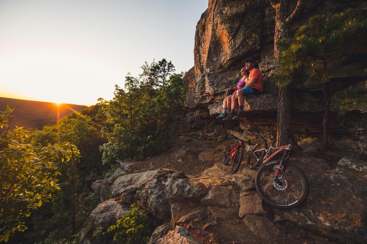 Cyclists resting on rocky cliff at sunset with bikes nearby.