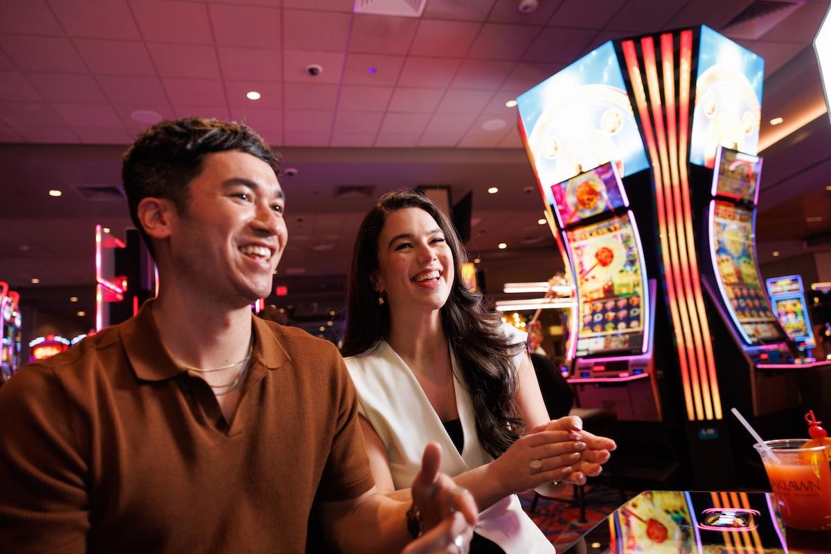 Casino scene with smiling couple at slot machines, bright colorful lights in background.
