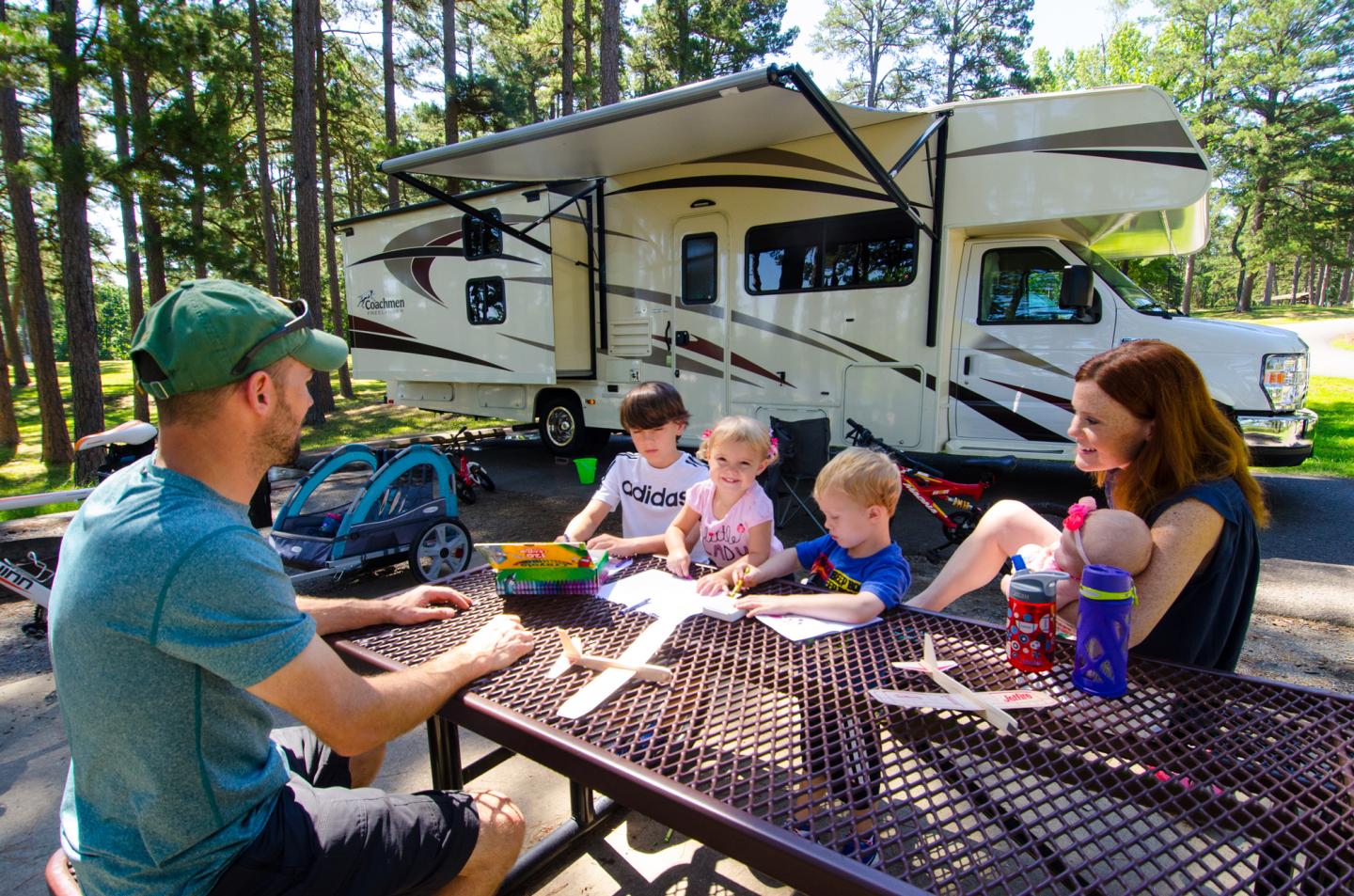 Family camping by an RV, enjoying a sunny outdoor meal at a picnic table.