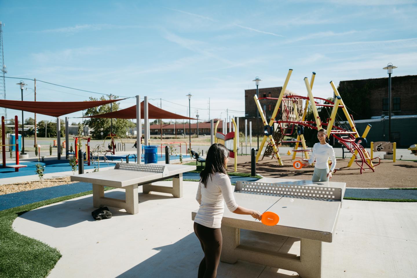 Two people play table tennis in a sunny playground with colorful equipment.