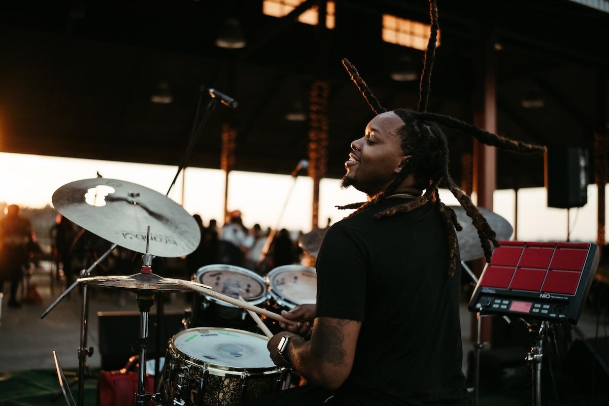 Drummer playing at sunset, with long dreadlocks and a focused expression.