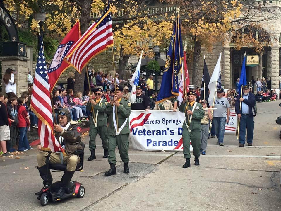Veterans parade with people holding flags, autumn trees in background.