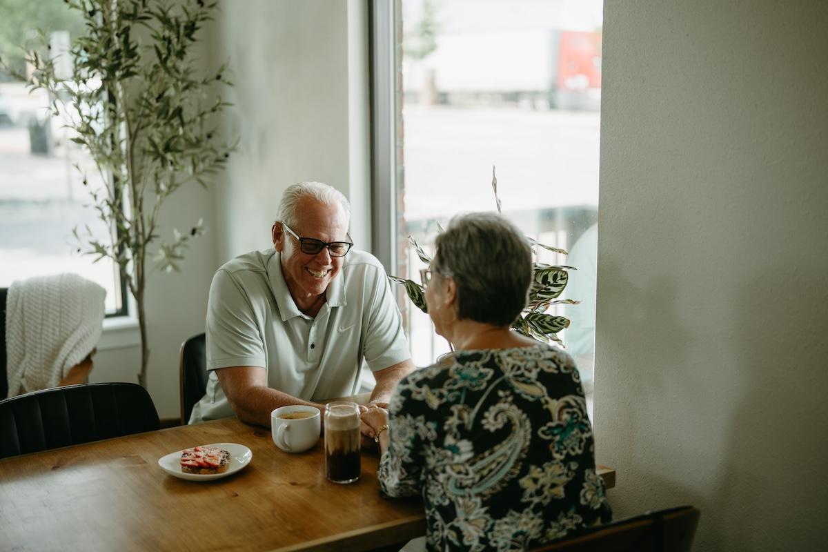 Elderly couple enjoying coffee and conversation at a cafe table near a window.