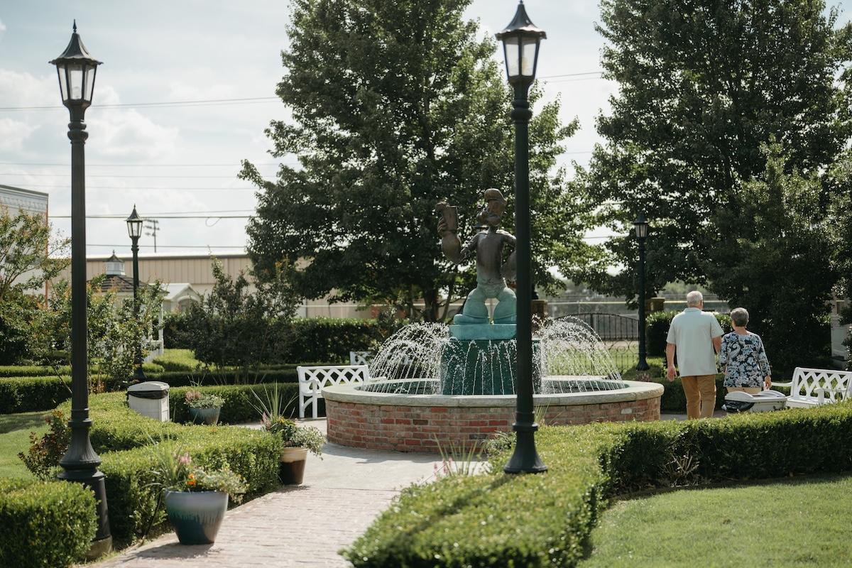 Pathway to fountain with benches, surrounded by green hedges and trees.