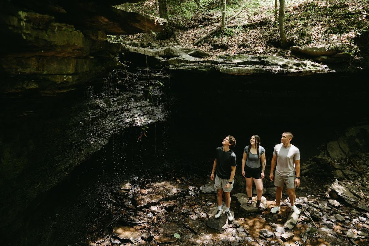Three people exploring a shaded rocky cave under dappled sunlight.