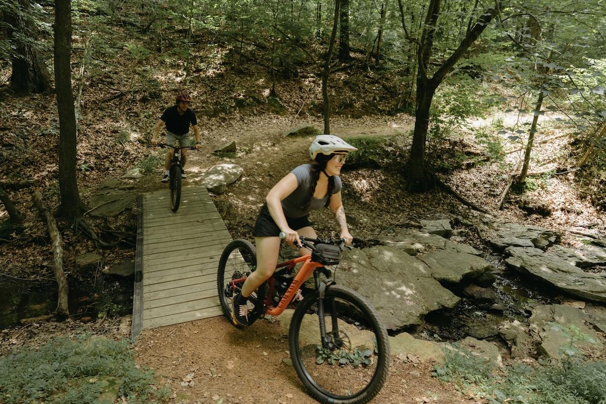 Two people mountain biking on a forest trail, crossing a small bridge.