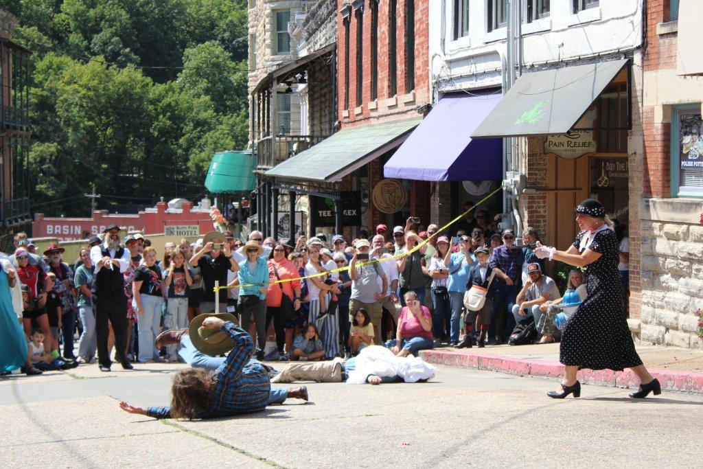 Street performance with actors and a crowd, woman pointing at person lying on the ground.