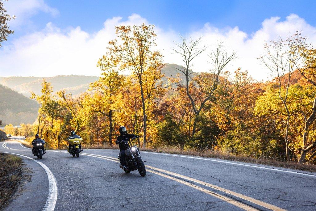 Motorcyclists on a scenic road with autumn trees and mountains.