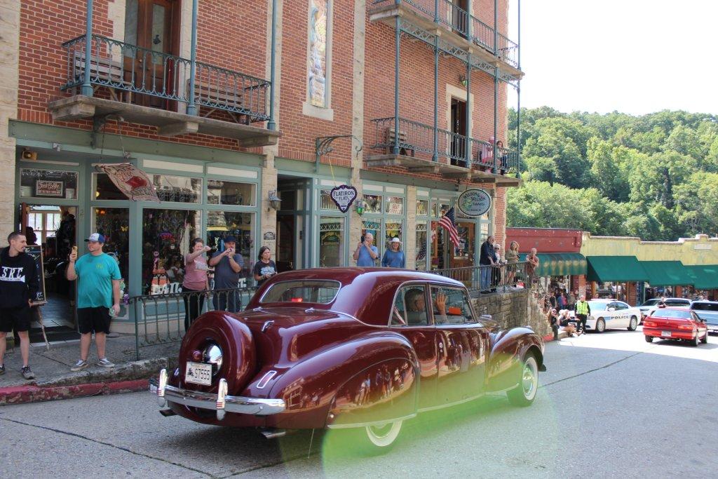 Classic maroon car on a street with onlookers and historic brick buildings.