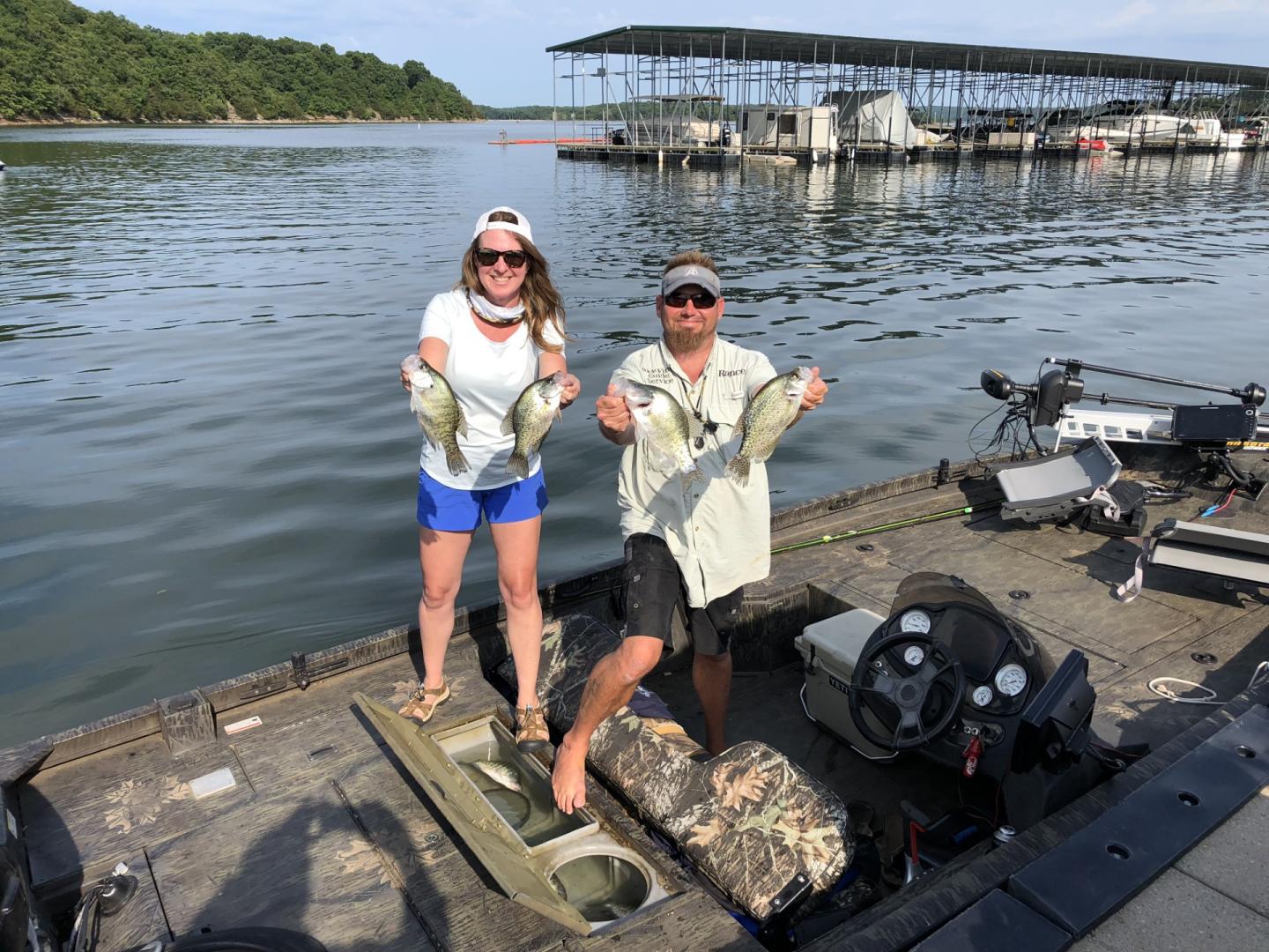 Two people on a boat holding fish, with a lakeside dock in the background.