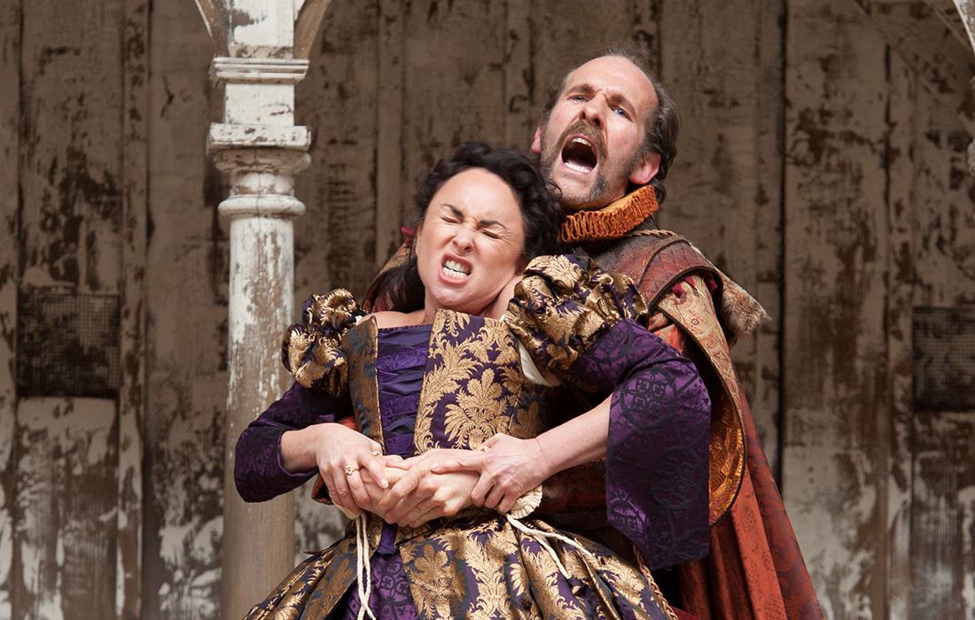 Two actors in ornate costumes, one restraining the other with a pained expression.