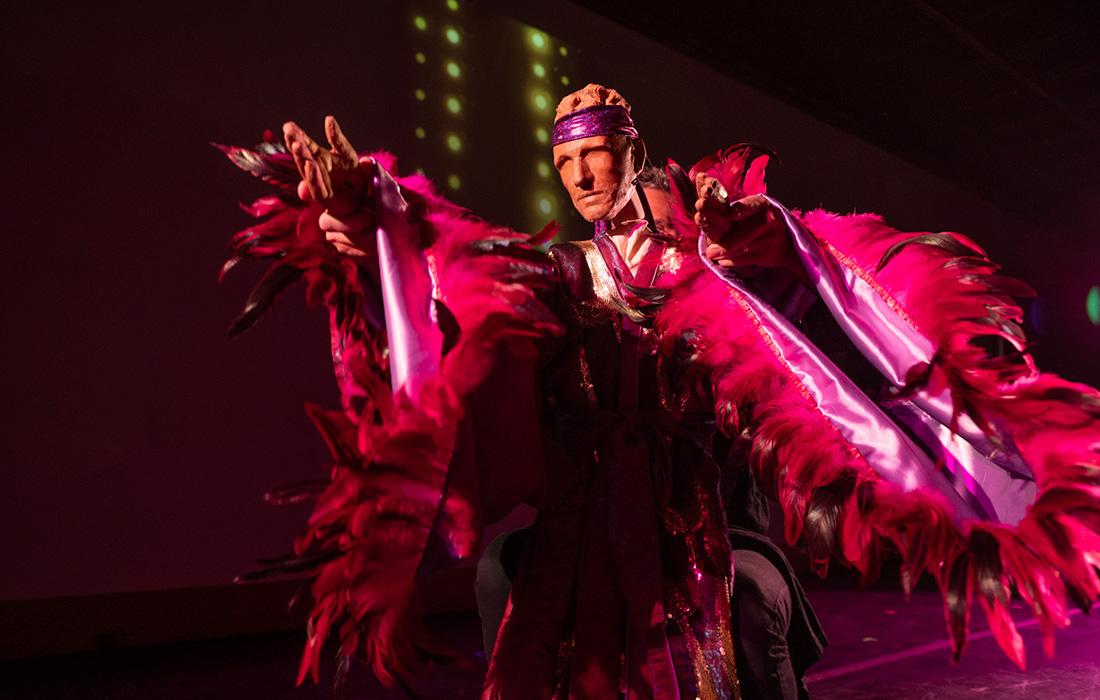 Man in vibrant feathered costume onstage, dramatic lighting.