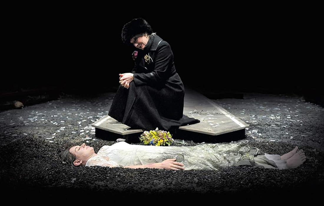 A person kneels by a tombstone, mourning, with a bouquet on a grave.