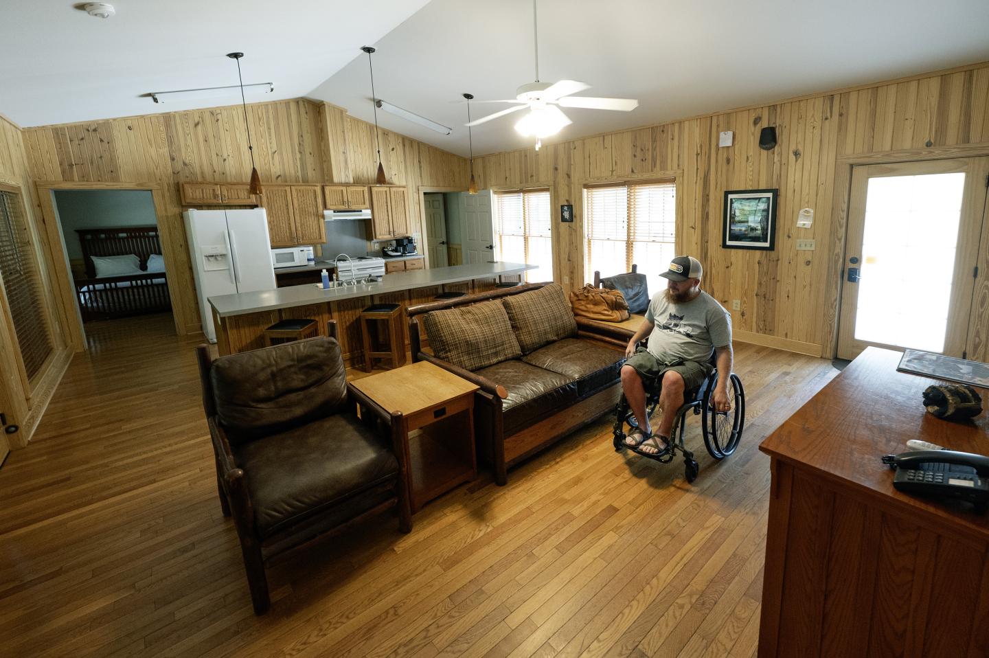 Cozy living room with a man in a wheelchair, wooden walls and floors, and ceiling fan.