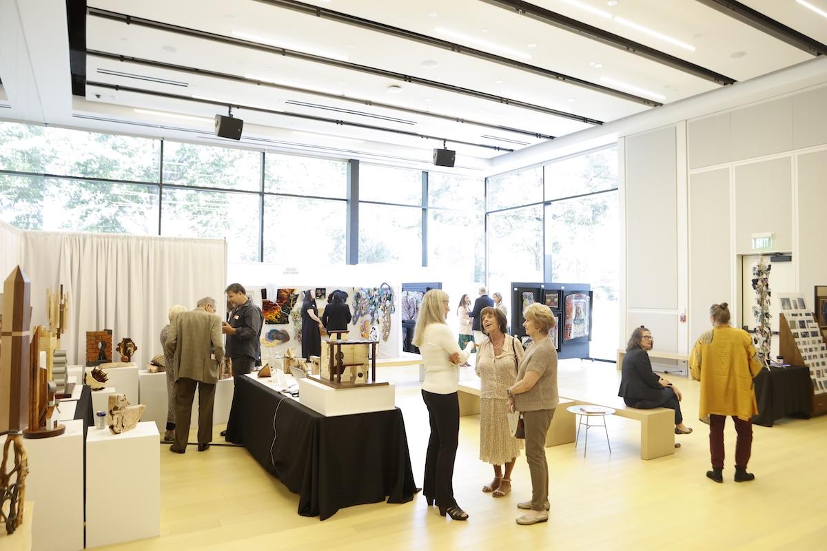 Art exhibit with people viewing and discussing artworks in a bright, spacious gallery.