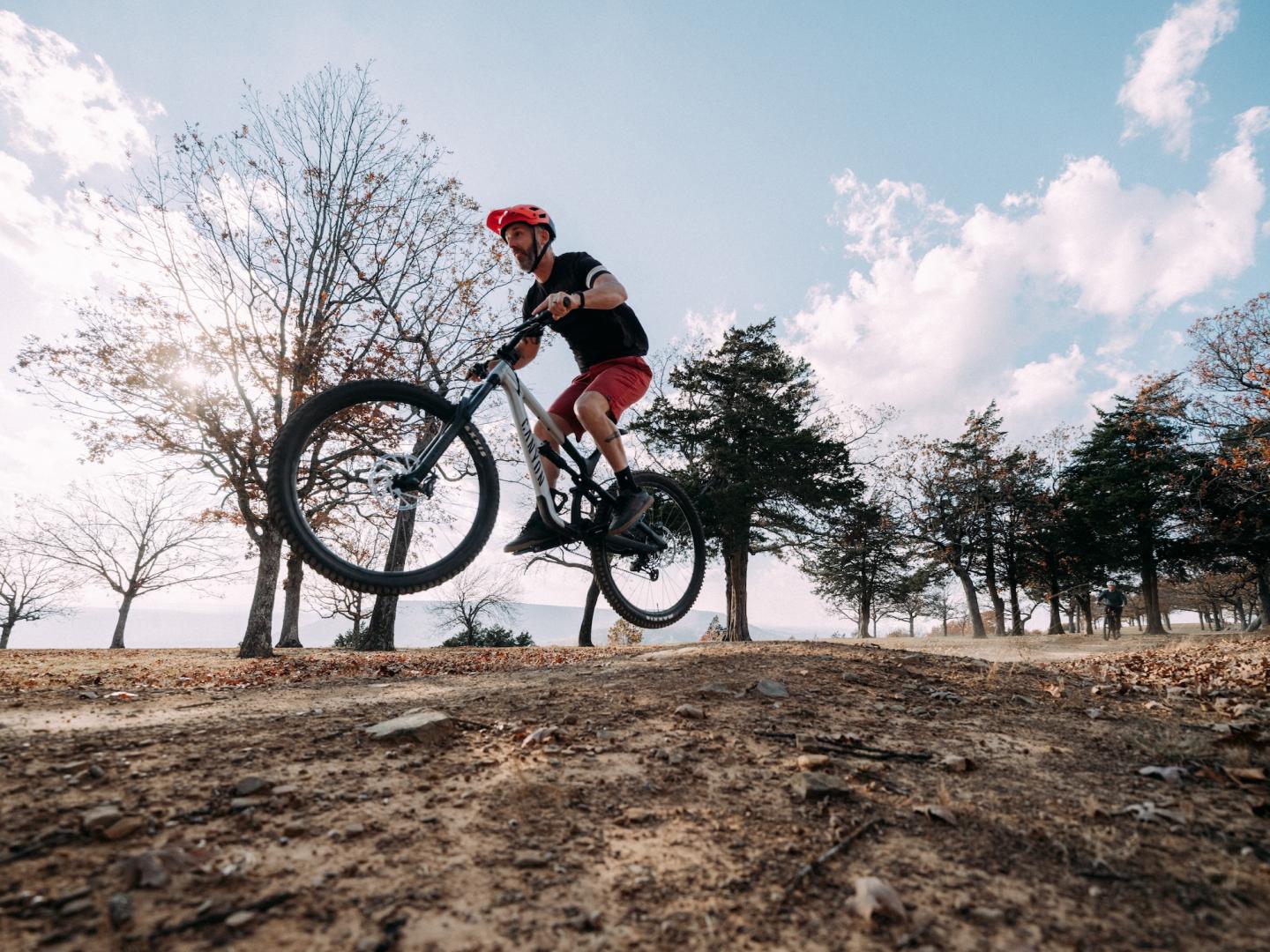 Cyclist jumps bike on dirt path, surrounded by trees and blue sky.