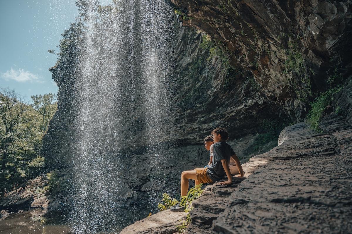 Person sitting near a waterfall on rocky ledge, sunny day.