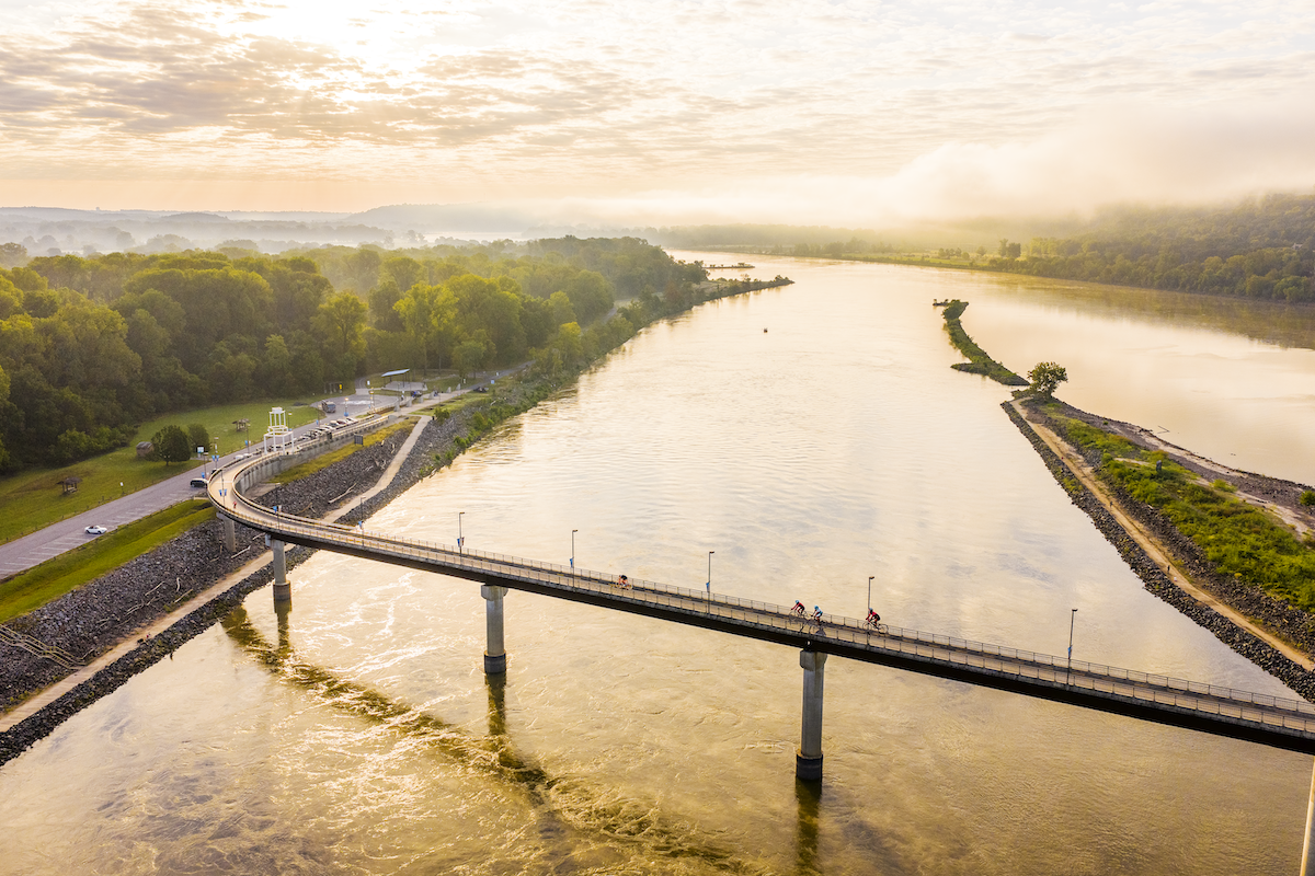 Aerial view of a bridge over a calm river at sunrise with green trees on the banks.