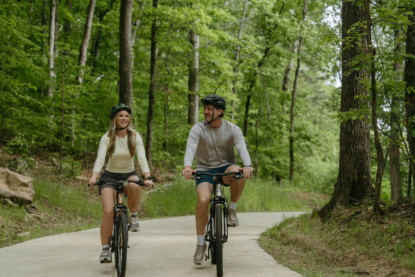 Two people cycling on a wooded trail, wearing helmets and casual clothes.