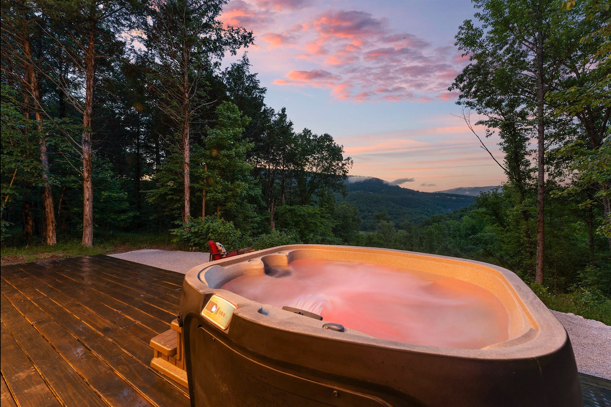 Hot tub on a wooden deck, sunset sky, surrounded by trees.