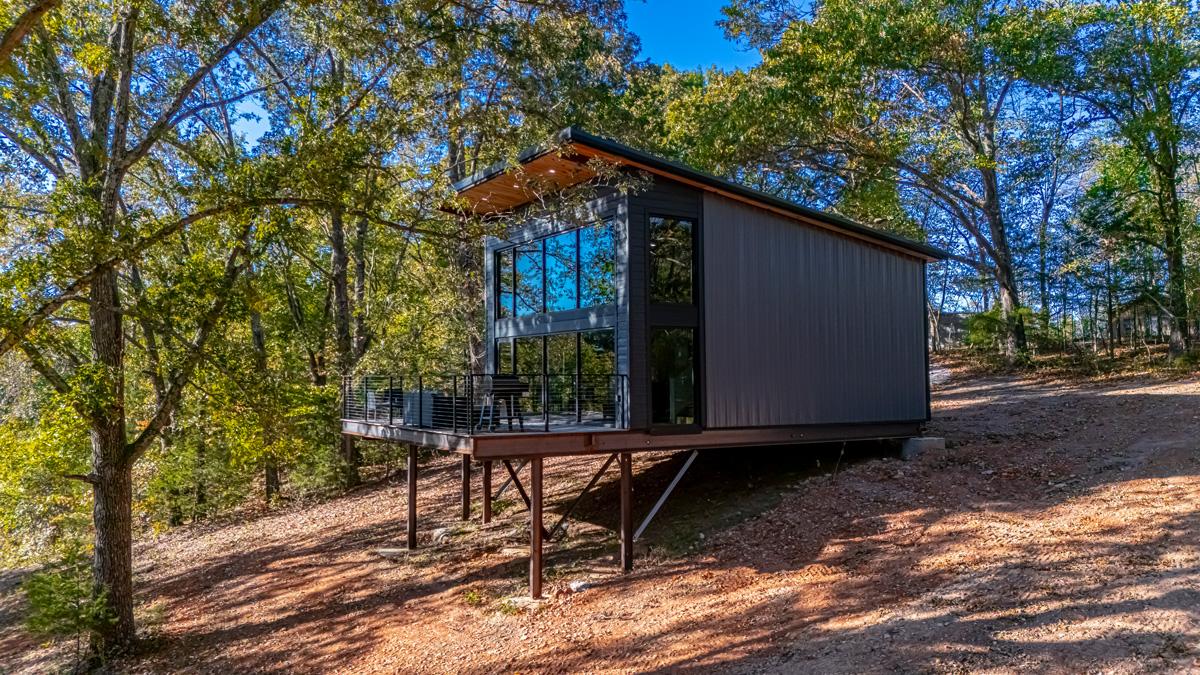 Modern cabin on stilts in a wooded area, under a clear blue sky.