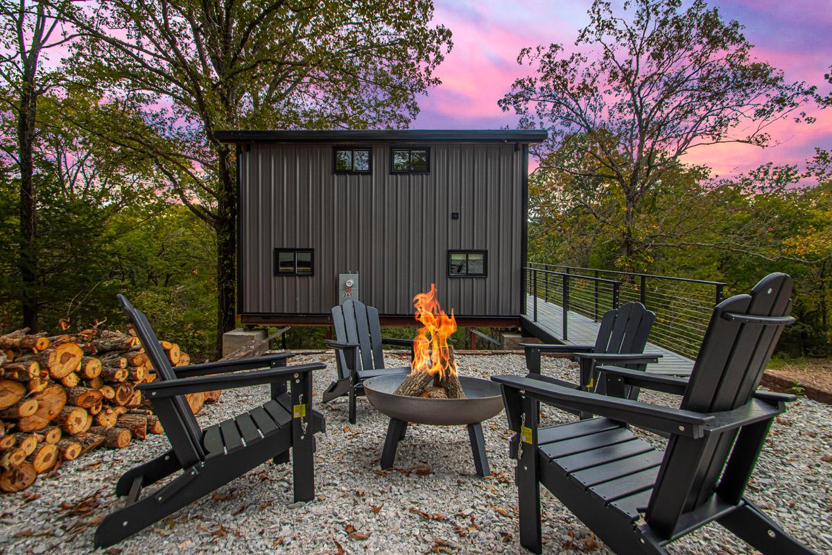 Modern cabin, fire pit, four chairs, sunset sky, and trees in the background.