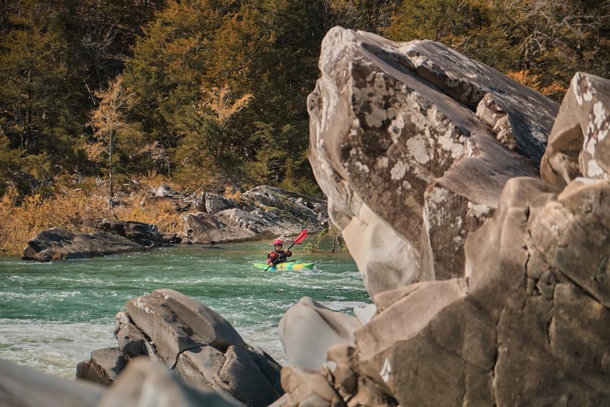 Kayaker navigating a rapid between large rocks amidst autumn trees.