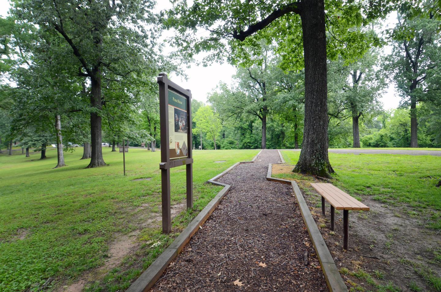 Park path with trees, a signpost, and a bench under a cloudy sky.