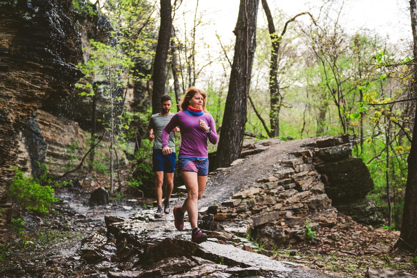 Two people jogging on a forest trail, surrounded by trees and rocky terrain.