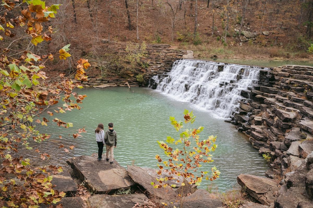 Two people stand by a waterfall in a forested area with autumn colors.