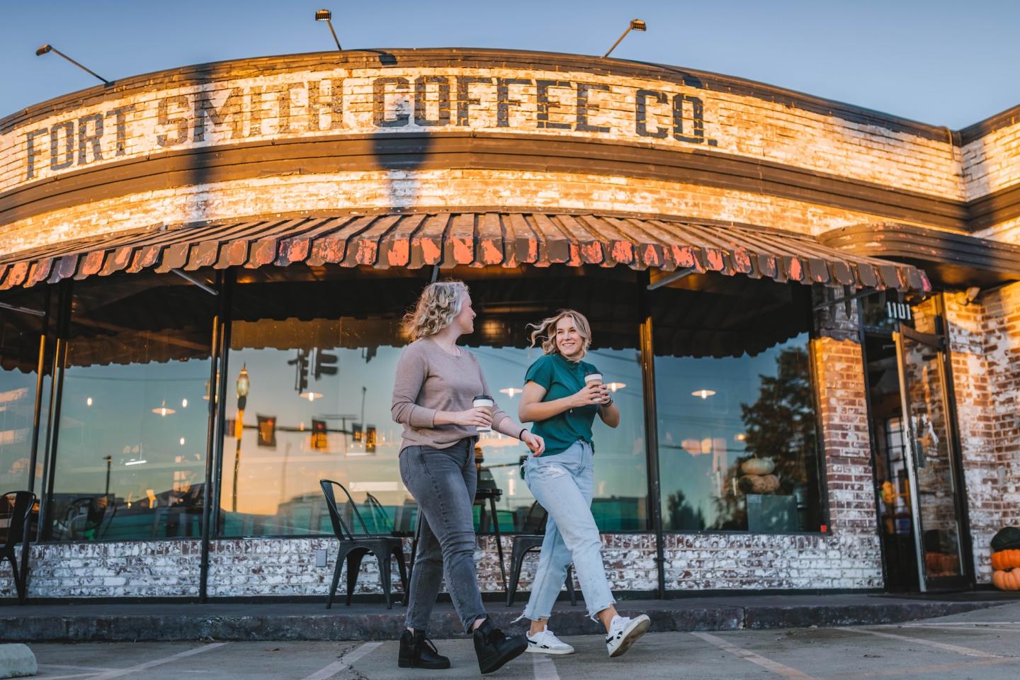 Two people walking with drinks in front of a cafe at sunset.