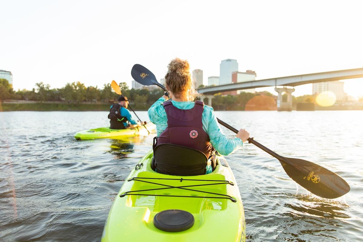 Kayakers on a river at sunset with bridge and city skyline.