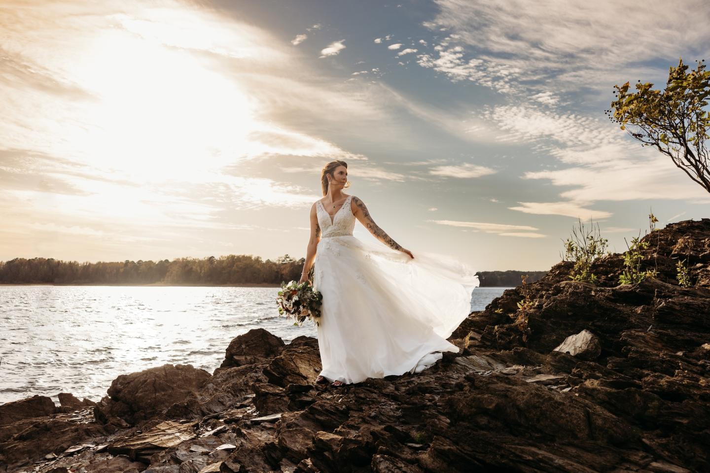 Bride in white gown stands on rocky shore, holding flowers, under a bright sky.