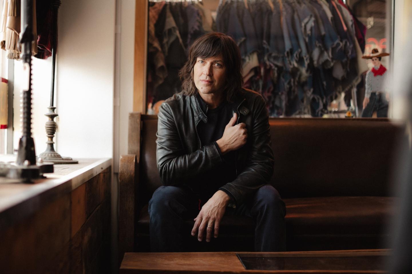 Person in a leather jacket sitting on a rustic bench inside a shop with hanging clothes.