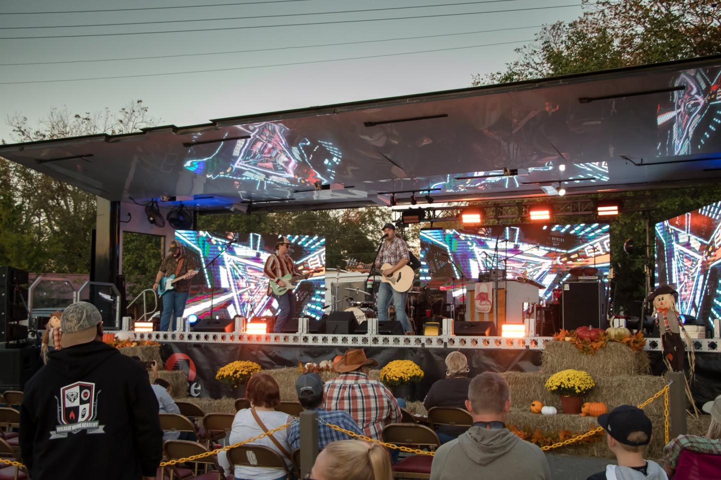 Outdoor concert with a band playing on stage, colorful lights, and seated audience.