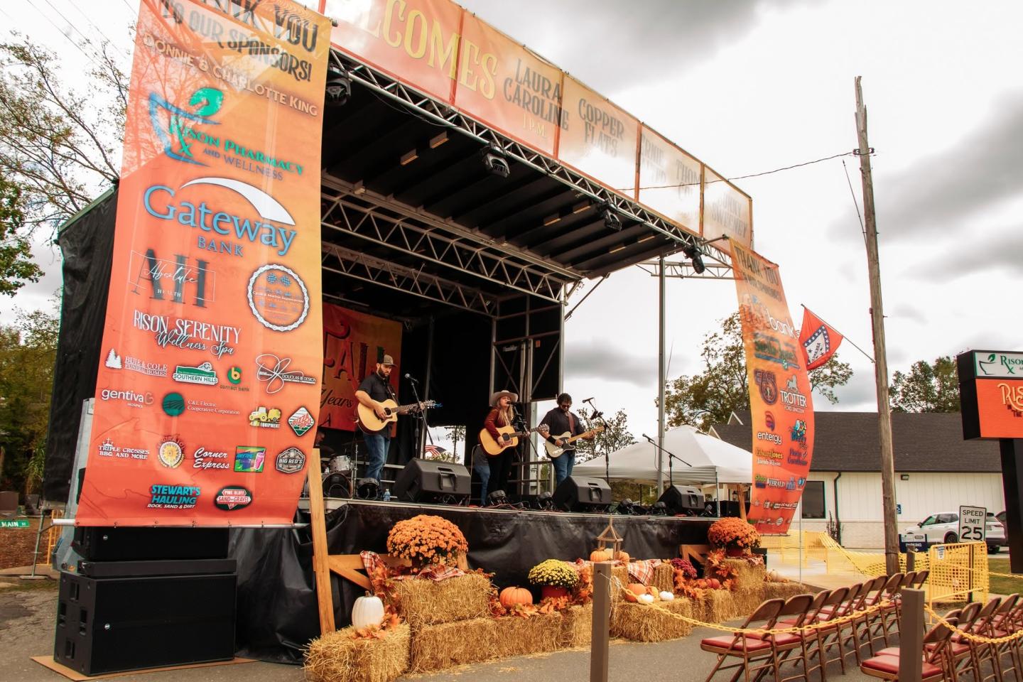 Outdoor concert stage with band performing, surrounded by pumpkins and hay bales.