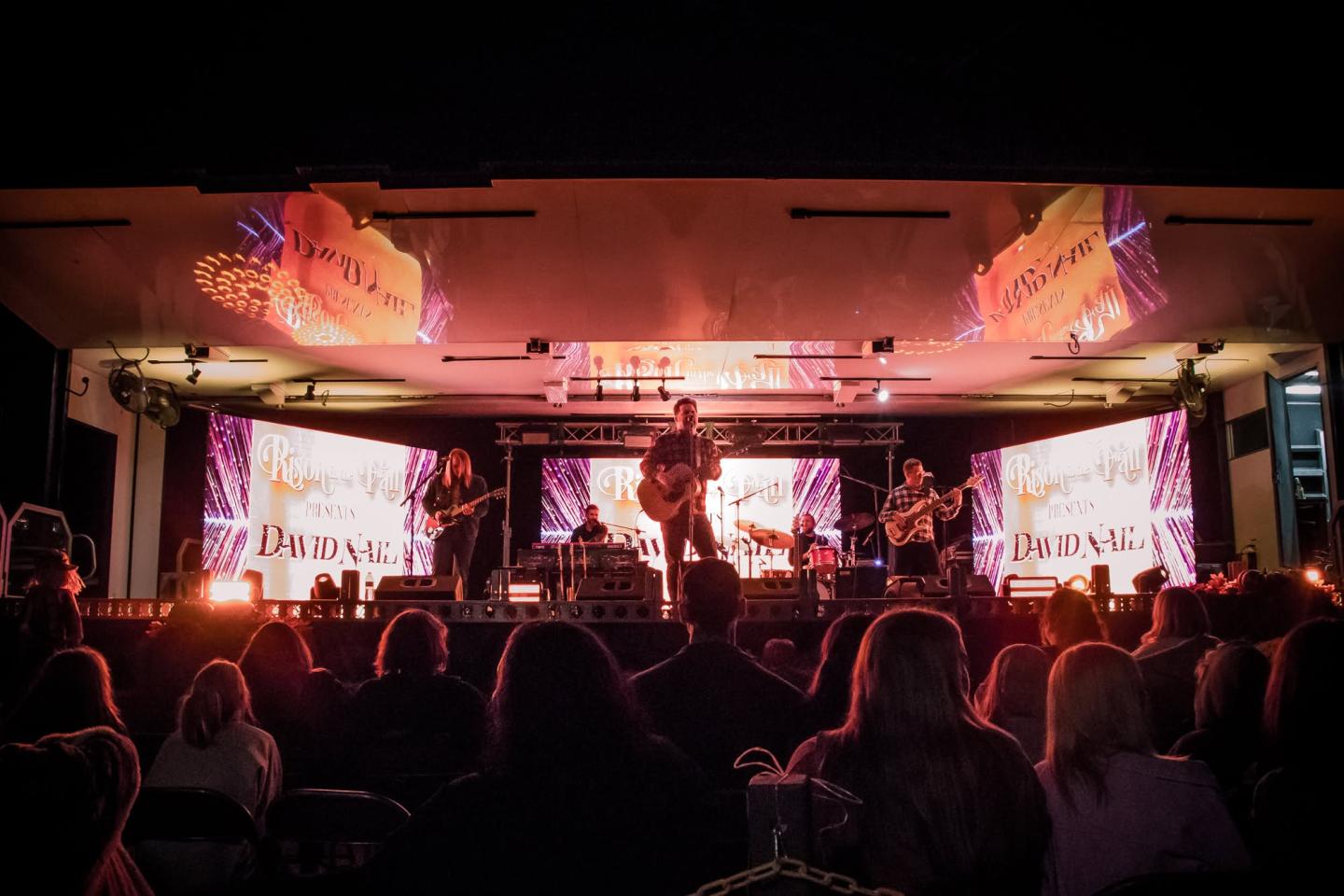Concert stage with musicians and colorful lights, audience silhouetted in foreground.