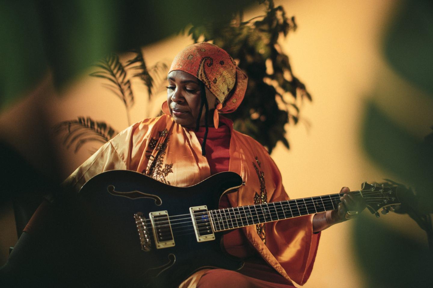 Musician in orange robes plays guitar amid lush plants.