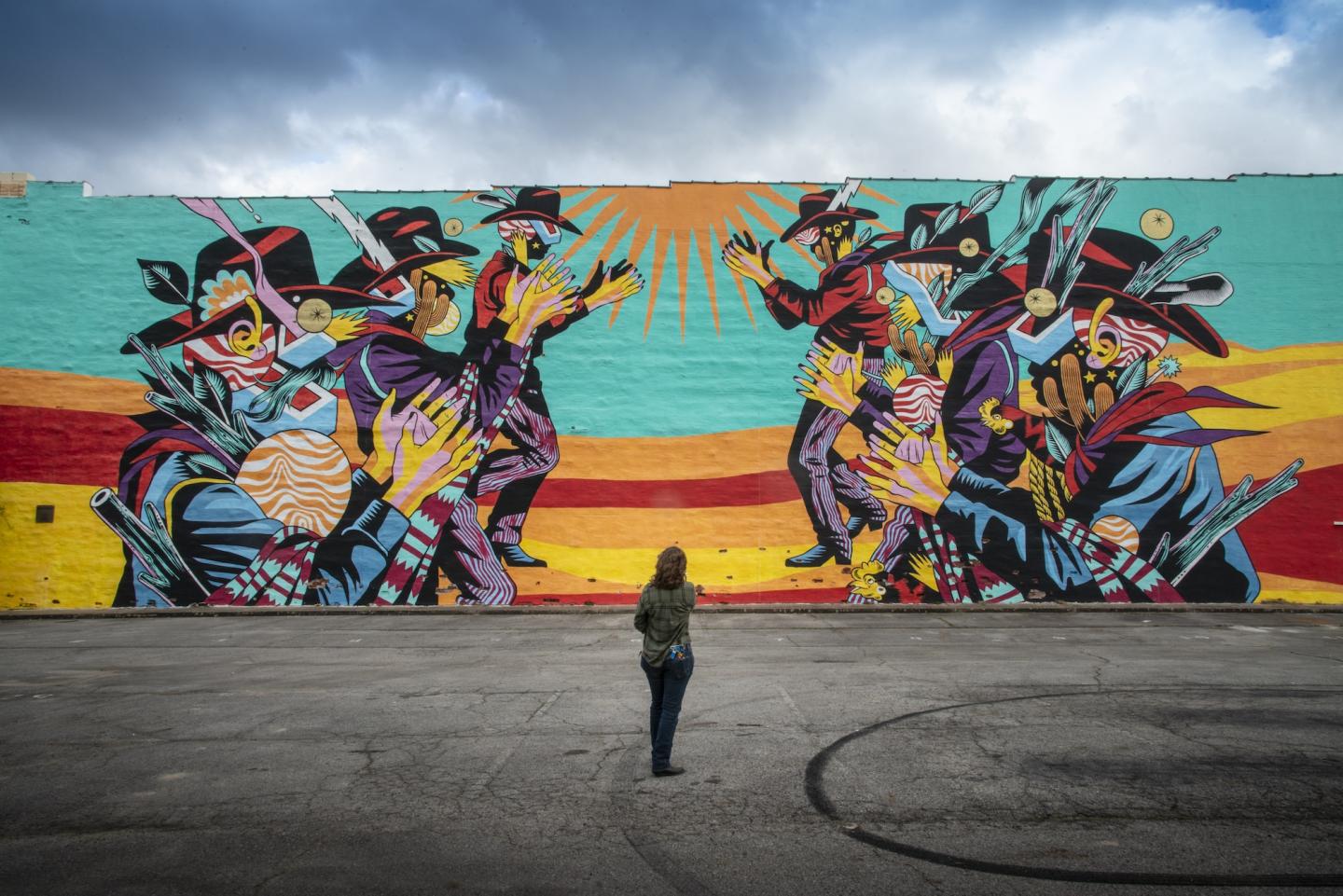 Colorful mural with figures and a person viewing it under a cloudy sky.