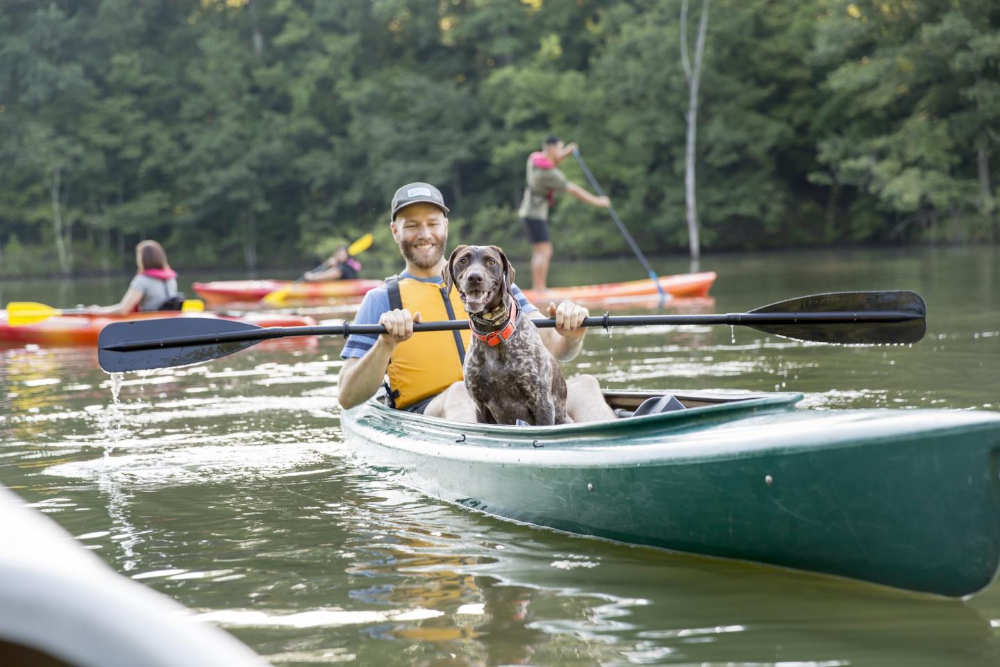 Man kayaking with a dog on a calm lake, others in kayaks nearby, surrounded by trees.