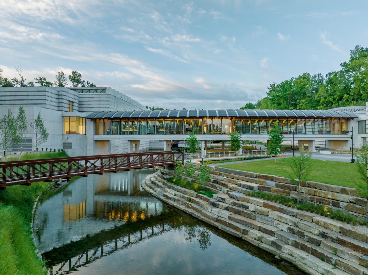Modern building with glass walls, arched roof, over a calm stream and landscaped garden.