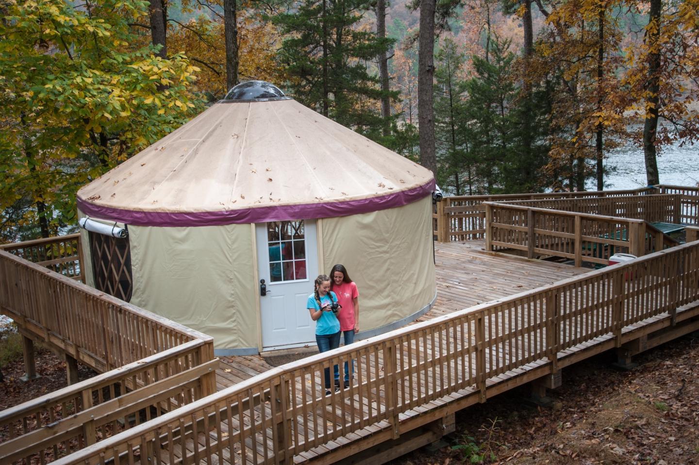 A yurt on a wooden deck in a forested area, with two people standing in front.