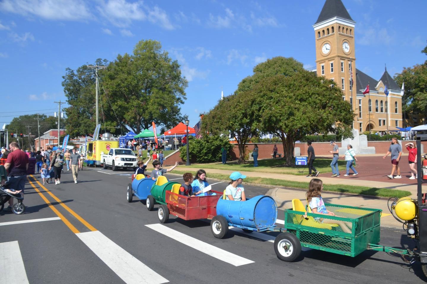 Children ride colorful toy train cars in a street parade near a clock tower.