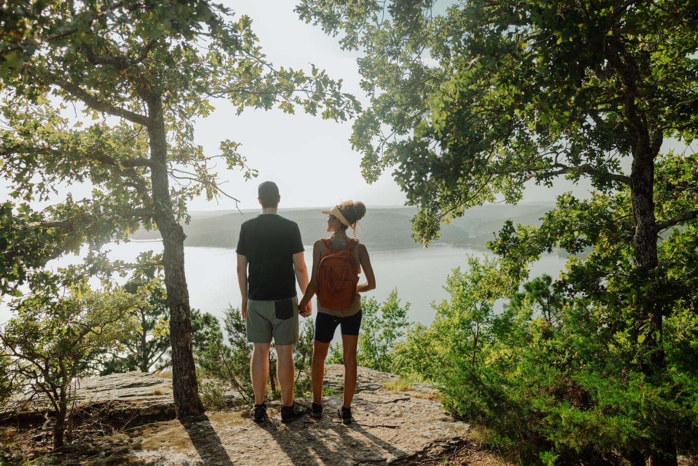 Couple standing on a hill, overlooking a lake surrounded by trees.