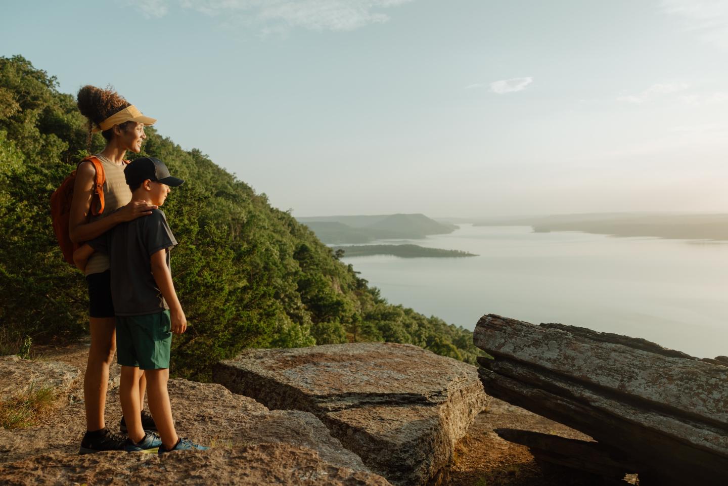 Two people stand on a rocky cliff, overlooking a misty lake and forested hills.