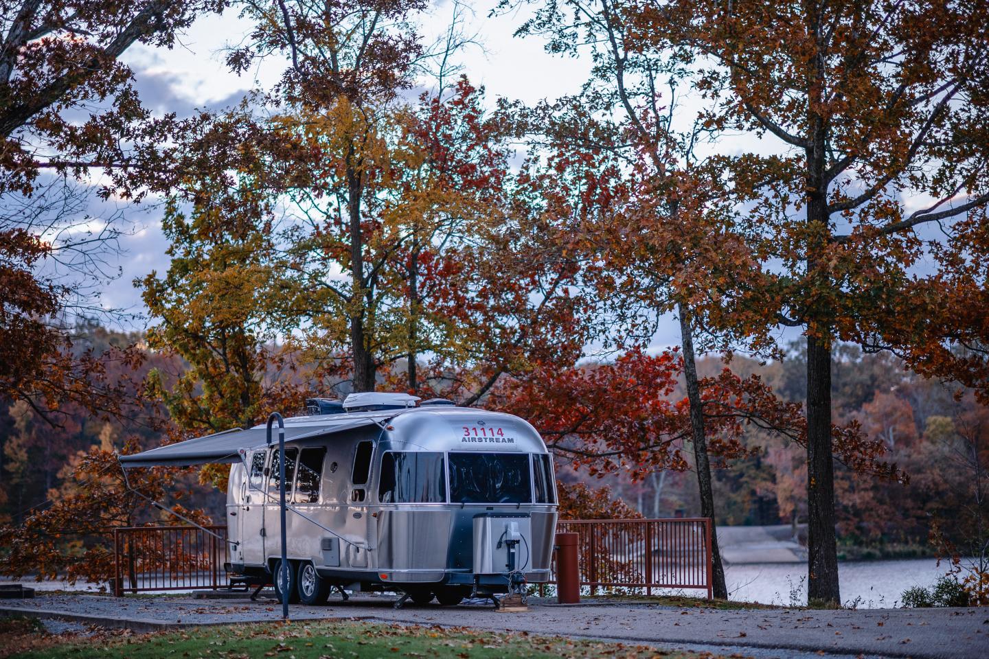 Silver Airstream trailer parked beneath autumn trees by a lake.