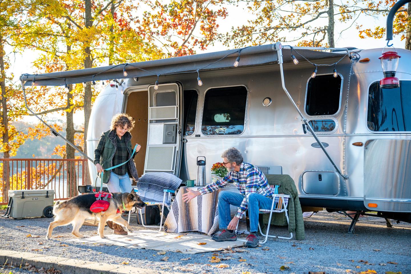 Couple with a dog relaxes outside a parked camper in a scenic, autumnal setting.